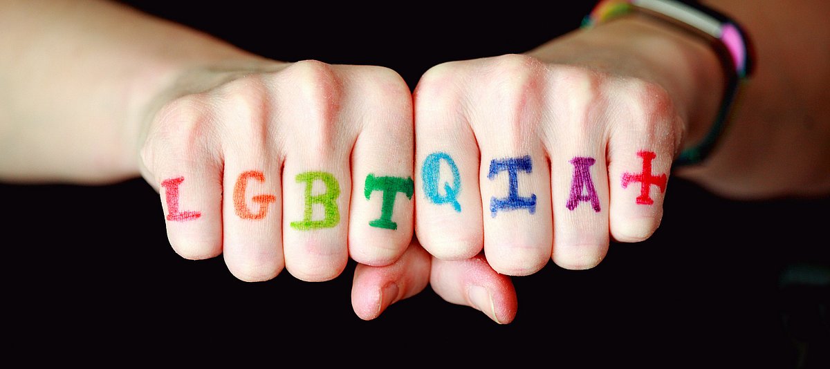 Das Bild zeigt zwei Fäuste. Über die Finger ist „LGBTQIA+“ in bunten Farben geschrieben