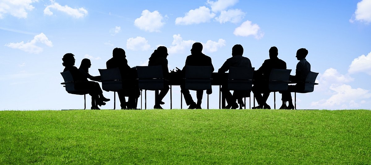 Eine Gruppe von Menschen sitzt an einem Besprechungstisch mitten auf einer grünen Wiese mit blauem Himmel