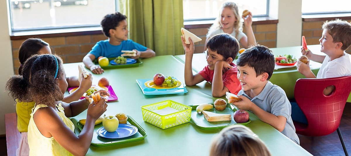 An zwei Tischen sitzen mehrere Kinder und essen Brot, Obst und Muffins
