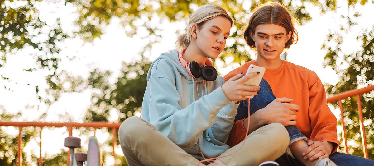 Zwei Jugendliche sitzen in einem Park mit Skateboard und schauen auf ein Smartphone