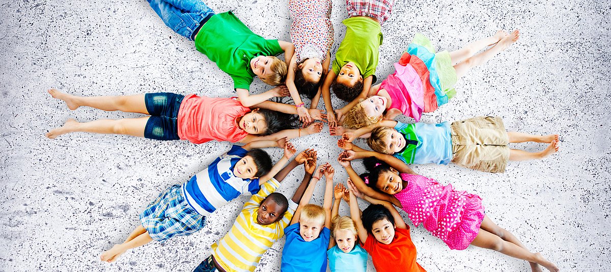 Kinder unterschiedlicher Herkunft liegen sternförmig an den Händen gefasst auf dem Boden