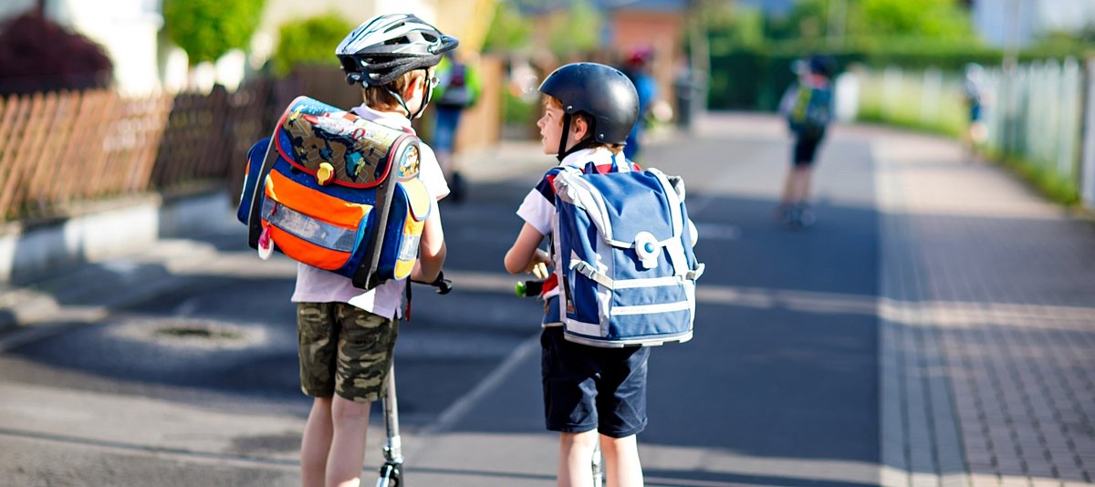 Zwei Jungen mit Schulranzen und Helmen auf fahren mit ihren Rollern auf einer Straße