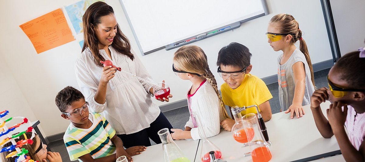Mehrere Kinder mit Schutzbrillen stehen um einen Tisch mit Reagenzgläsern und beobachten eine erwachsene Person mit einem Reagenzglas in der Hand