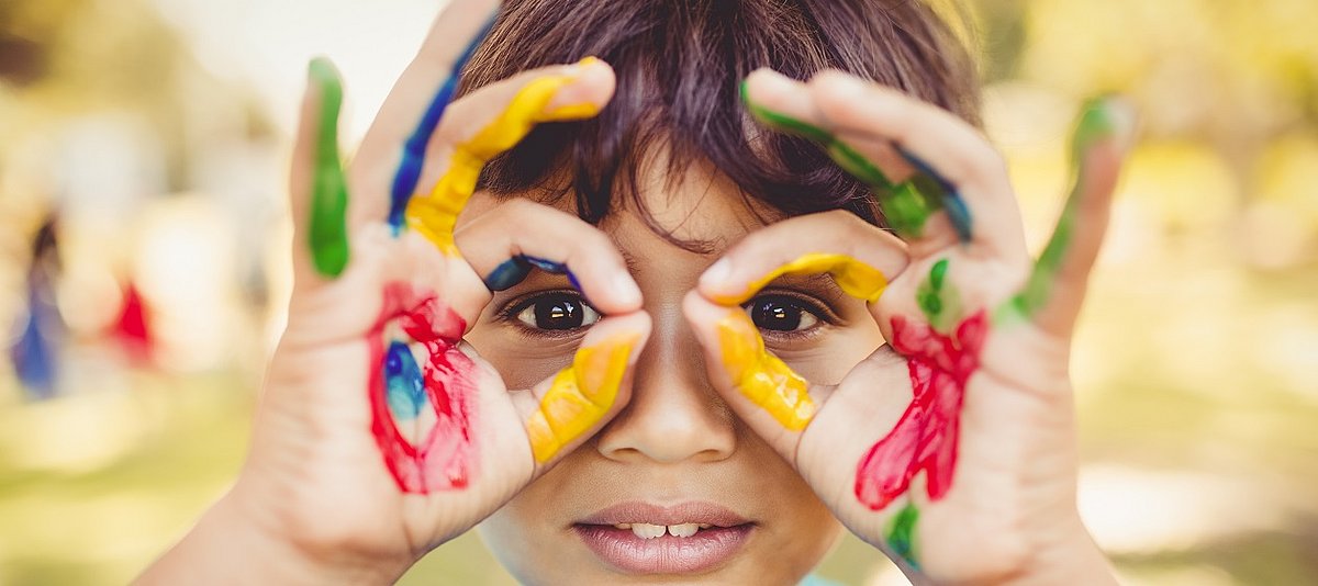 Ein Kind mit bunt bemalten Händen formt eine Brille aus den Fingern und schaut hindurch