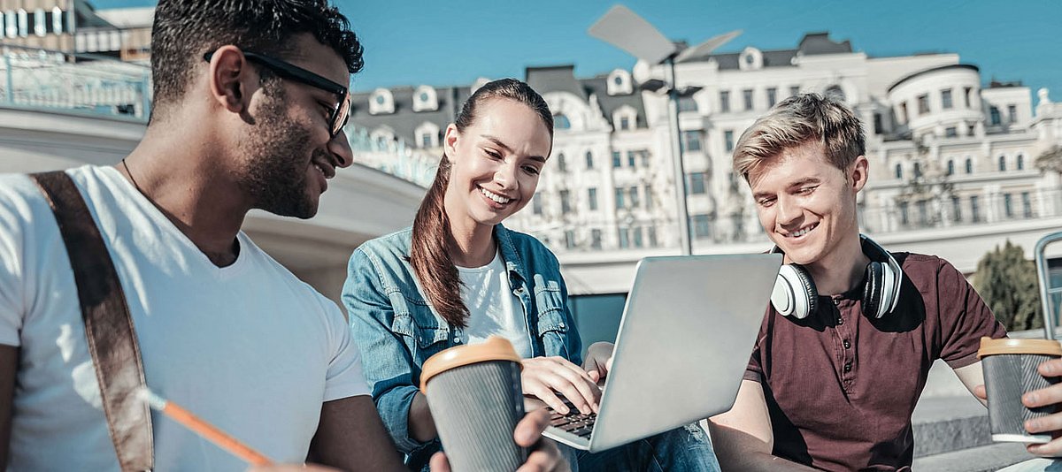 Drei Jugendliche unterschiedlicher Herkunft sitzen lächelnd auf einer Treppe und schauen gemeinsam auf einen Laptop.