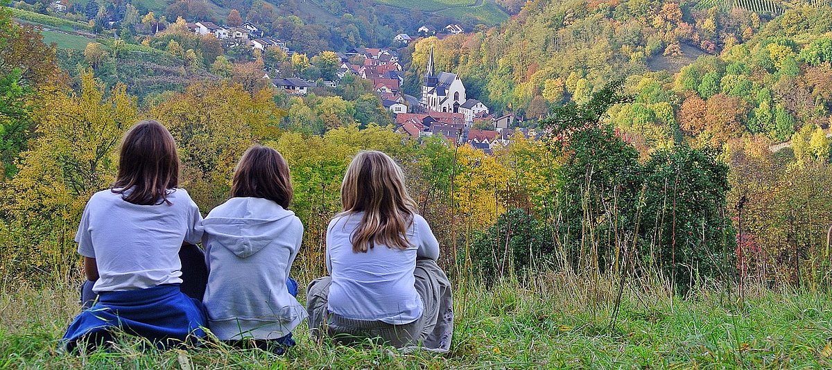 Drei Kinder sitzen mit dem Rücken zur Kamera und schauen von einer Anhöhe auf ein Dorf
