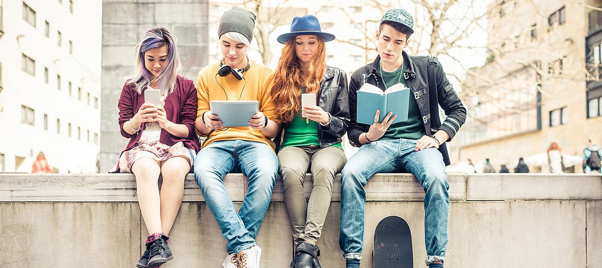 Vier Jugendliche sitzen auf einer Mauer, jeder hält ein Medium in der Hand, darunter zwei Smartphones, ein Tablet und ein Buchn 