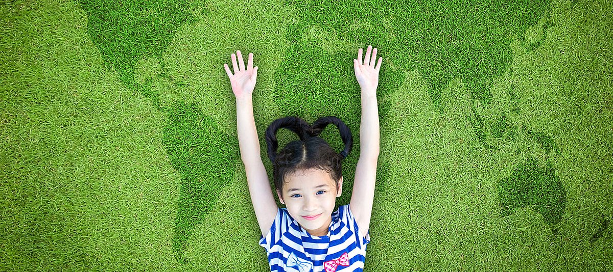 Ein Mädchen im Grundschulalter liegt auf einer Weltkarte aus Rasen und lächelt.