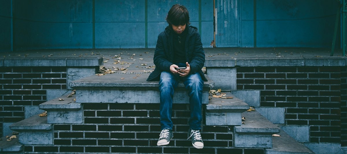 Junge sitzt allein auf einem Treppenabsatz und schaut auf sein Smartphone