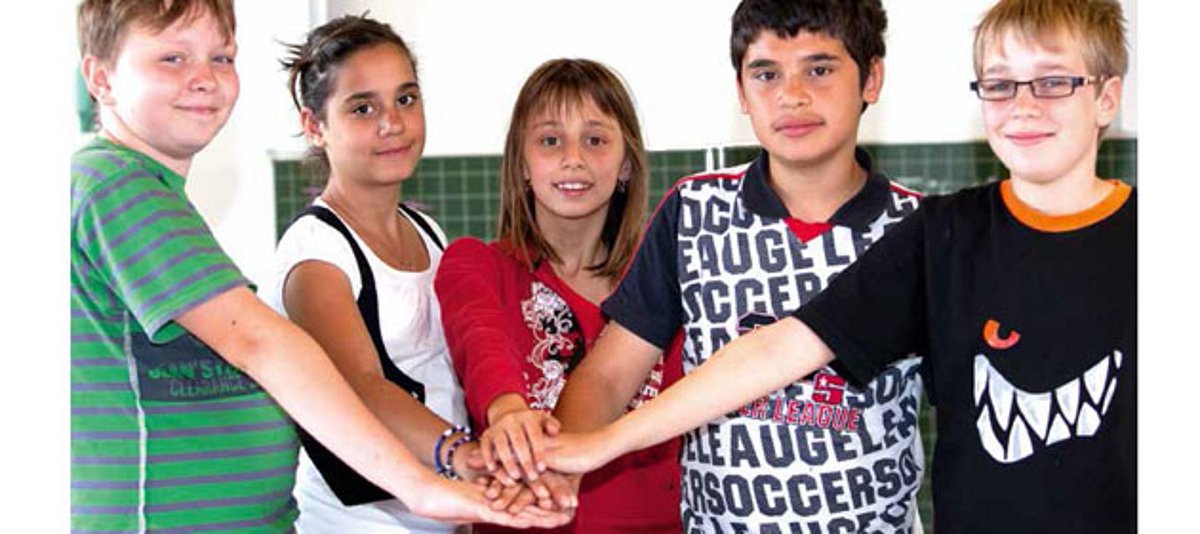 Fünf Kinder stehen mit  übereinandergelegten Händen vor einer Tafel im Klassenzimmer