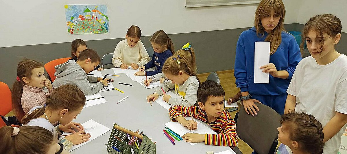 Kinder malen an einem Tisch und sprechen mit jungen Freiwilligen 