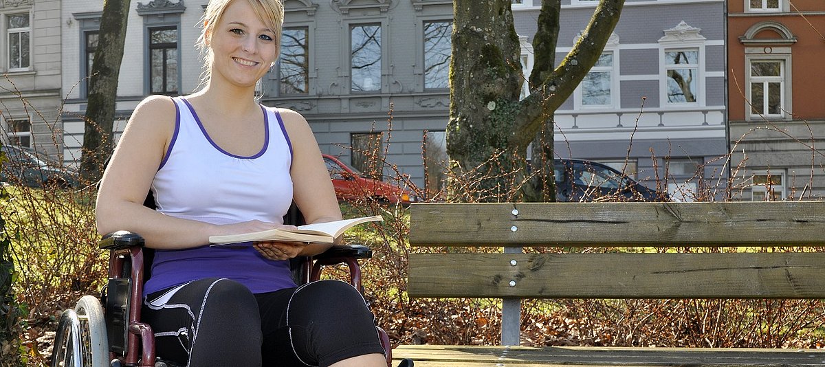 ein Mädchen im Rollstuhl sitzt lächelnd neben einer Parkbank