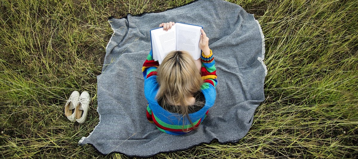 Eine junge Frau sitzt auf einer Decke in einer Wiese und liest in einem Buch