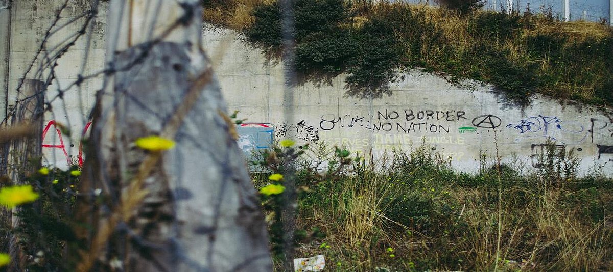 Es ist eine Mauer zu sehen, auf der in Graffiti der Schriftzug „No Borders, No Nations“ zu lesen ist. 