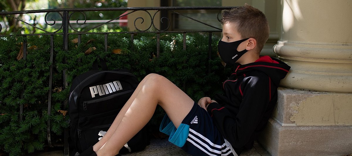Ein Junge mit Mund-Nase-Bedeckung sitzt mit seiner Schultasche auf einer Mauer