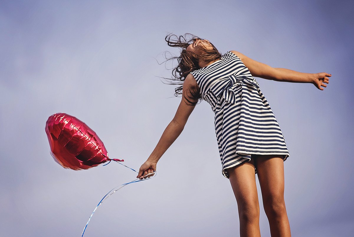 Ein Mädchen in gestreiftem Kleid und mit rotem, luftgefülltem Aluminiumballon in Herzform macht unter heiterem Himmel einen Sprung.