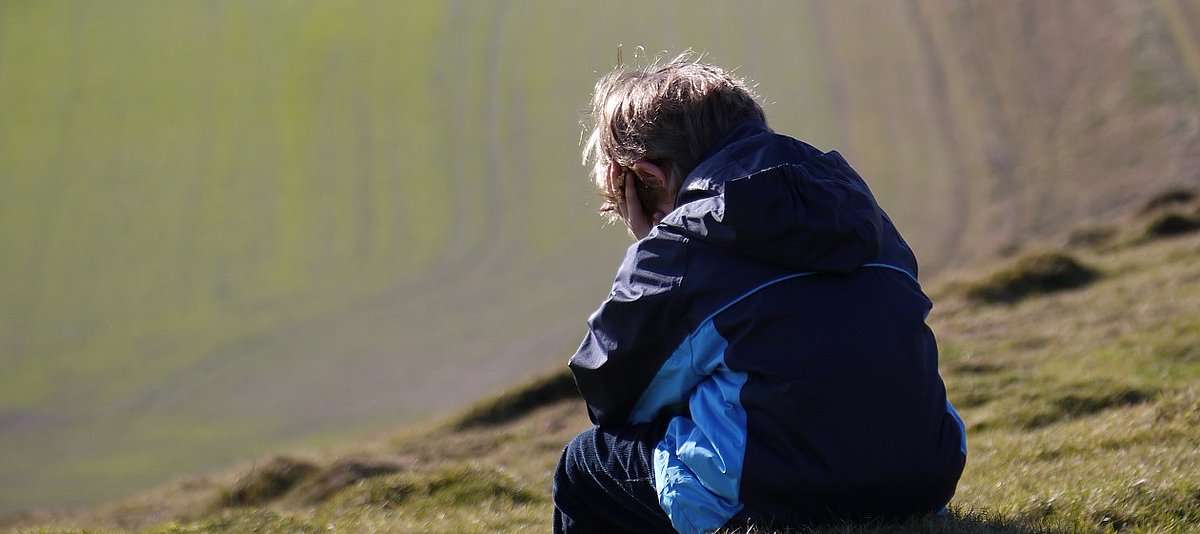 Ein Kind von hinten fotografiert sitzt auf einer Wiese in den Bergen und hat den Kopf in die Hände gestützt