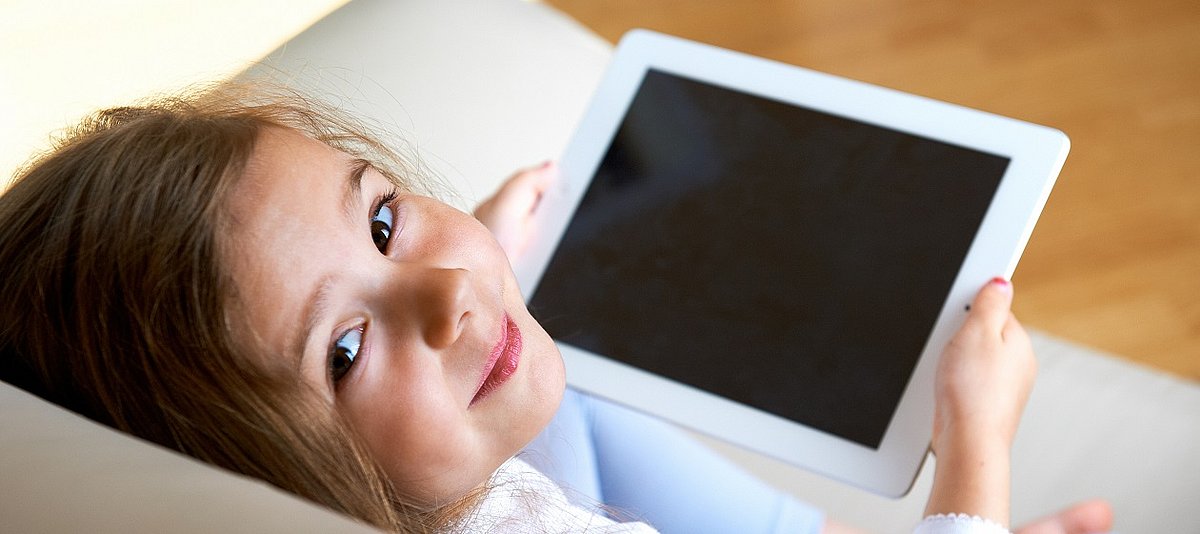 Ein Kind sitzt auf dem Sofa und hält ein Tablet in der Hand, der Bildschirm ist schwarz