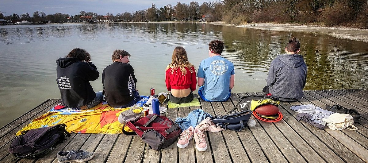 Fünf Jugendliche sitzen im Vorfrühling auf einem Bootssteg eines Sees.