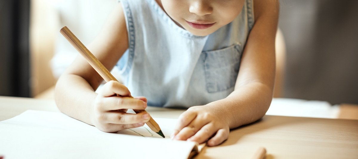 Kind sitzt an einem Tisch und schreibt etwas mit einem großen Bleistift