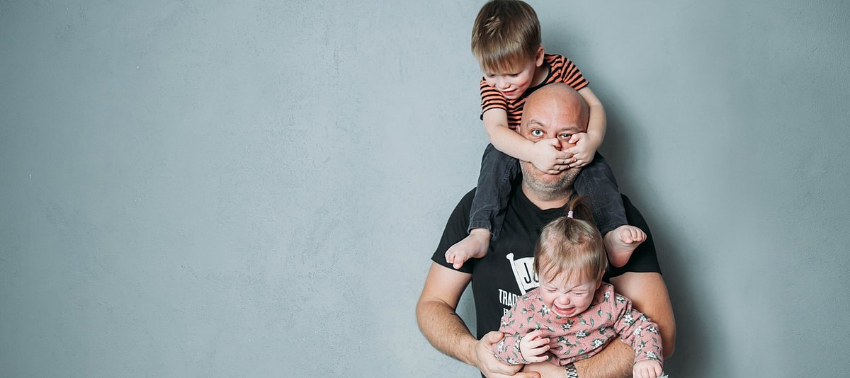 Ein getrsster Vater trägt zwei Kinder, das ältere sitze auf seinen Schultern und hält sich unglücklich an seinem Kopf fest