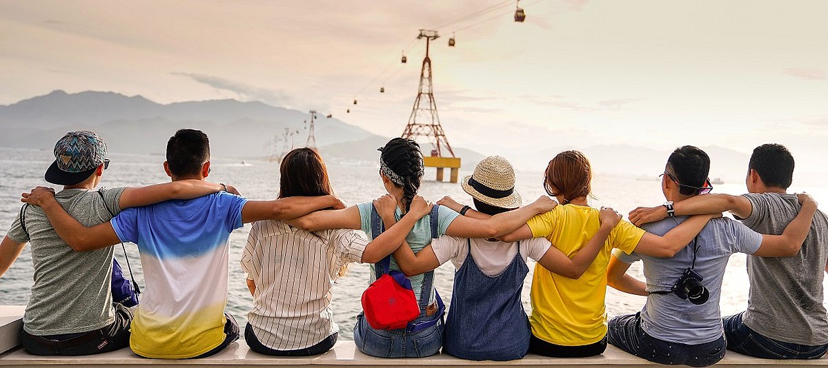 Eine Gruppe Jugendlicher von hinten fotografiert sitzt Arm in Arm und schaut auf einen See, über den eine Seilbahn gespannt ist 
