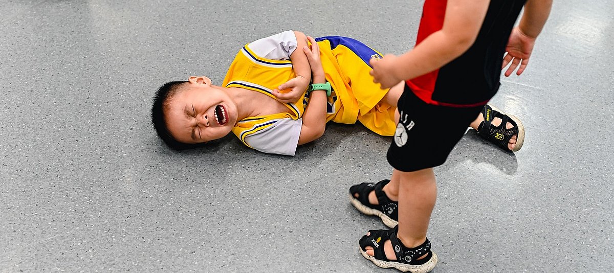 Ein Kind in Sprotkleidung liegt mit schmerzverzerrtem Gesicht auf dem Boden und hält sich den Arm, ein anderes Kind steht daneben