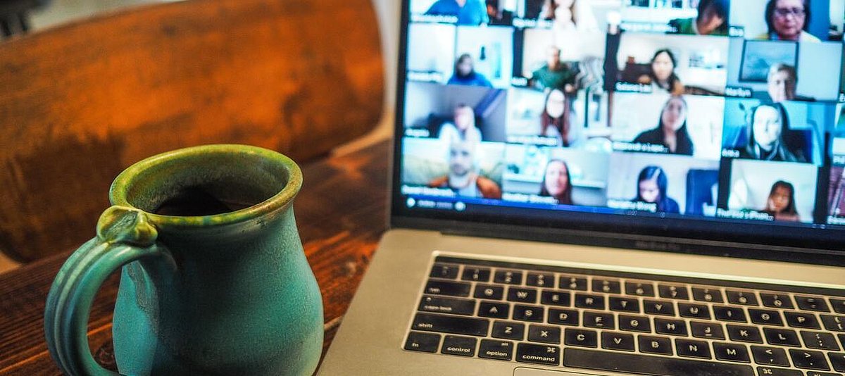 Eine Keramiktasse steht vor einem Laptop. Auf dessen Bildschirm läuft ein virtuelles Treffen mit vielen Teilnehmenden.