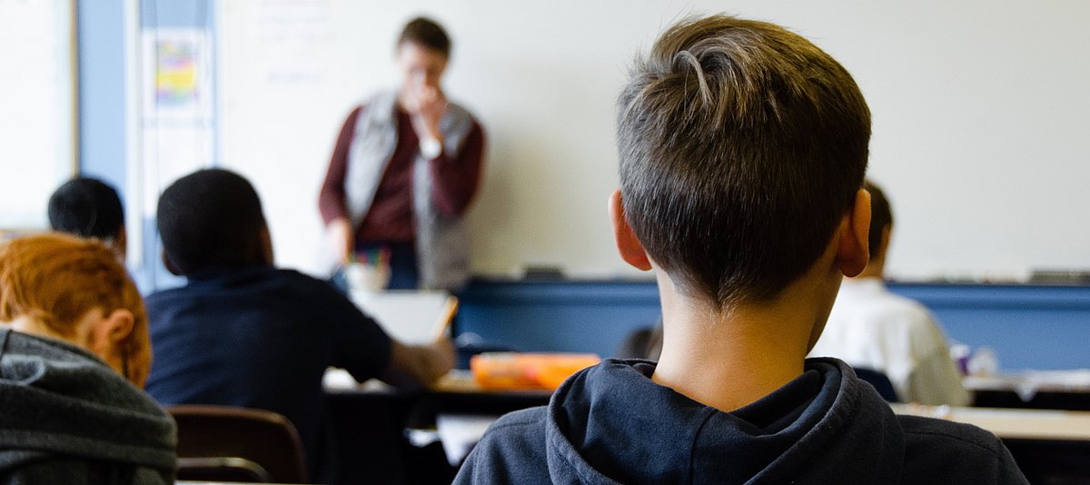 Jugendliche sitzen in einem Klassenraum, sie blicken zur Tafel und sind von hinten zu sehen