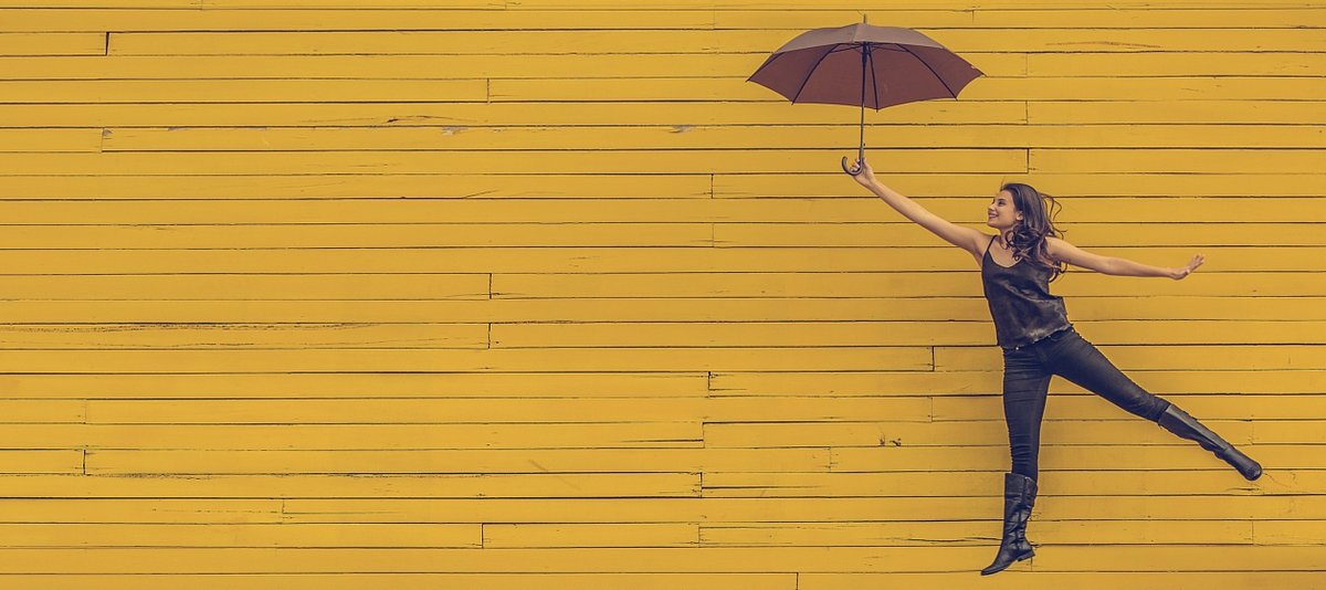 Eine Jugendliche springt schwebend mit einem Sonneschirm vor einer gelben Wand